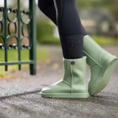 Jasnozielone, gumowe buty typu UGG na nogach w czarnych legginsach, obok zielonego ogrodzenia.