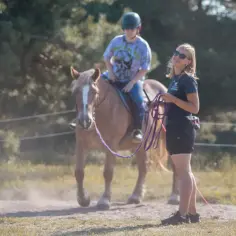 Dziewczyna w okularach prowadzi jazdę na lonży. Na koniu siedzi młody jeździec w kasku.